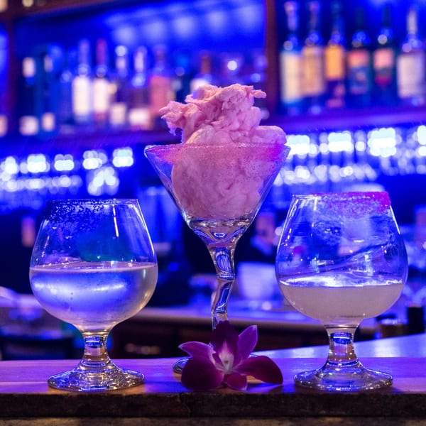 Three cocktails, one with pink cotton candy, on a bar with blue lighting and a purple orchid.