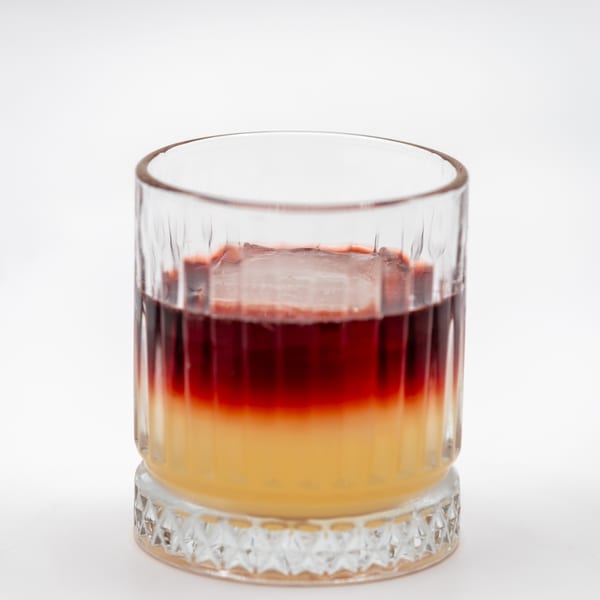 A vibrant, layered cocktail with yellow and red liquids, and a large ice cube, in a clear glass.