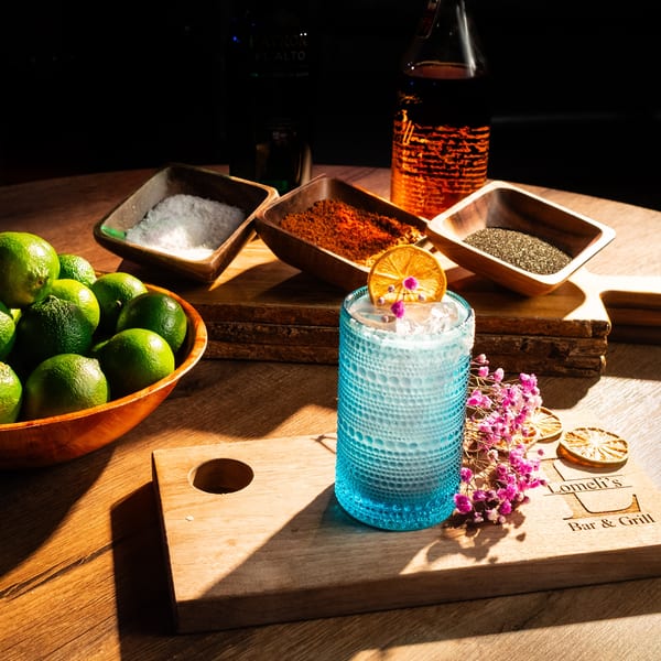 A blue cocktail with dried orange and flowers, surrounded by limes, salt, chili powder, and chia seeds.