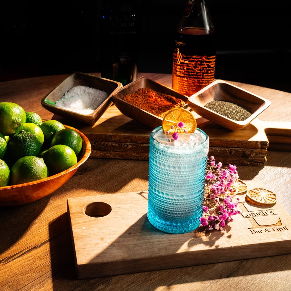 A blue cocktail with dried orange and flowers, surrounded by limes, salt, chili powder, and chia seeds.
