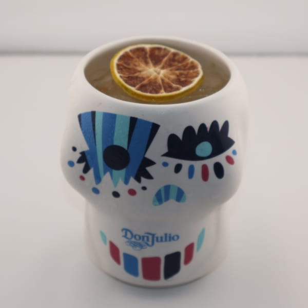 A cocktail in a white skull-shaped mug with colorful designs and a dried lime slice.