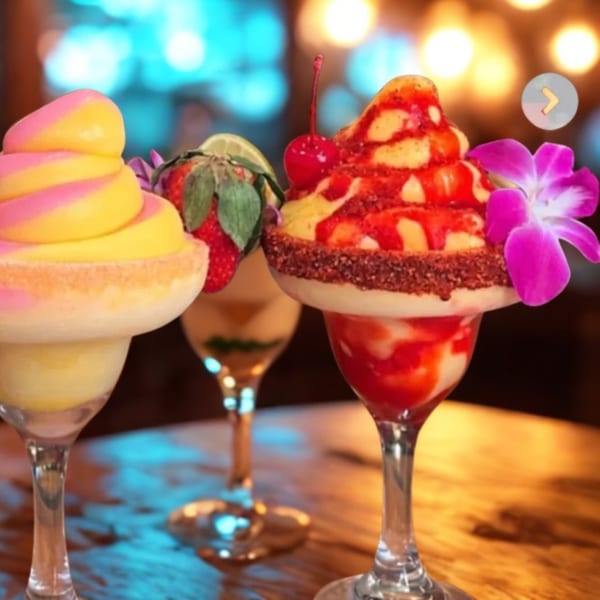 Three colorful frozen drinks with fruit and flower garnishes on a wooden table.