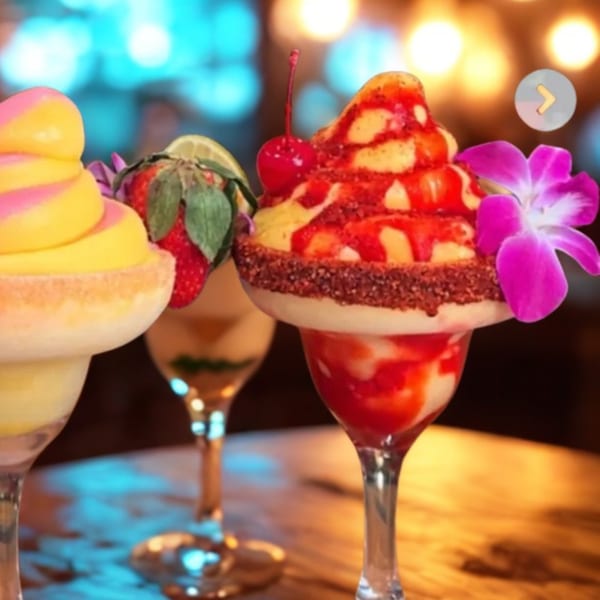 Three colorful frozen drinks, garnished with fruit and flowers, on a wooden table with blurred lights.