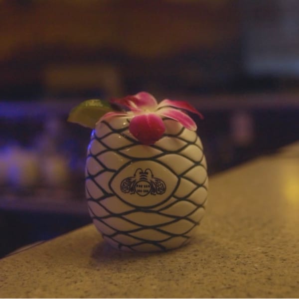 A pineapple-shaped cocktail glass with a lime wedge and pink flower on a bar.