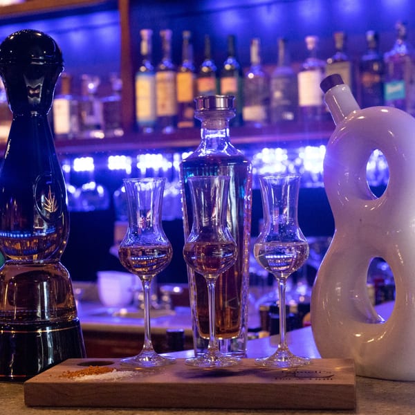 Three tequila glasses and bottles on a bar, with salt and spice on a wooden board.
