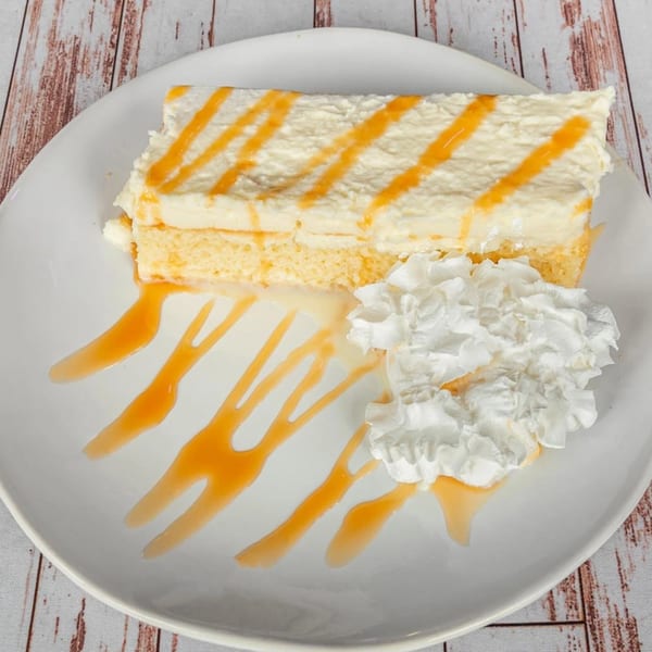 A slice of Tres Leches cake with caramel drizzle and whipped cream on a white plate.
