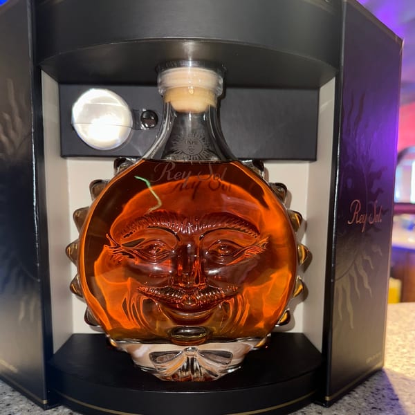 A bottle of Rey Sol tequila, shaped like a sun with a face, in its dark display box.