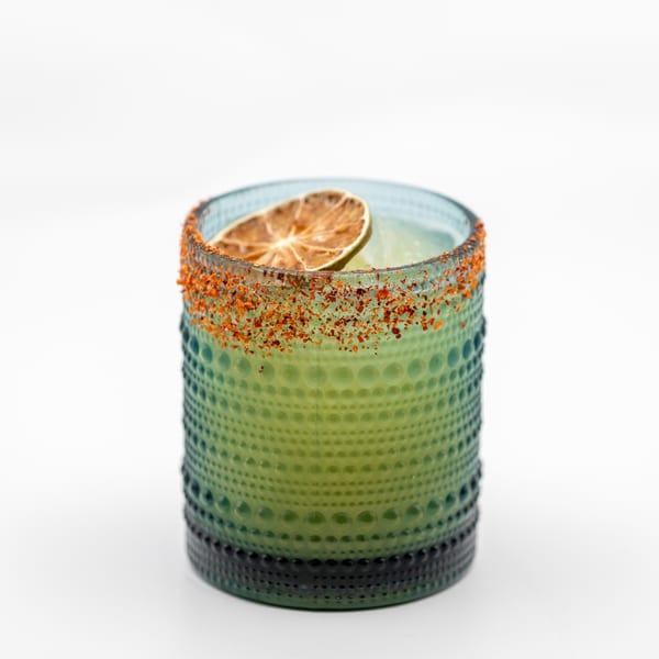 A vibrant green cocktail in a textured glass, rimmed with chili flakes and garnished with a dried lime slice.