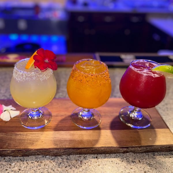 Three colorful Lomeli's Mexican Bar and Grill margaritas, garnished with fruit and flowers, on a wooden board.