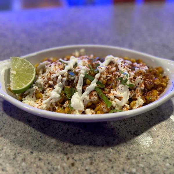 Mexican street corn (Esquites) with cotija cheese, crema, chili powder, cilantro, and a lime wedge.