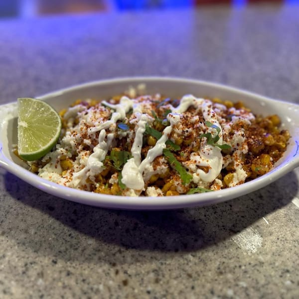 Mexican street corn in a white dish, topped with cheese, crema, chili powder, and a lime wedge.