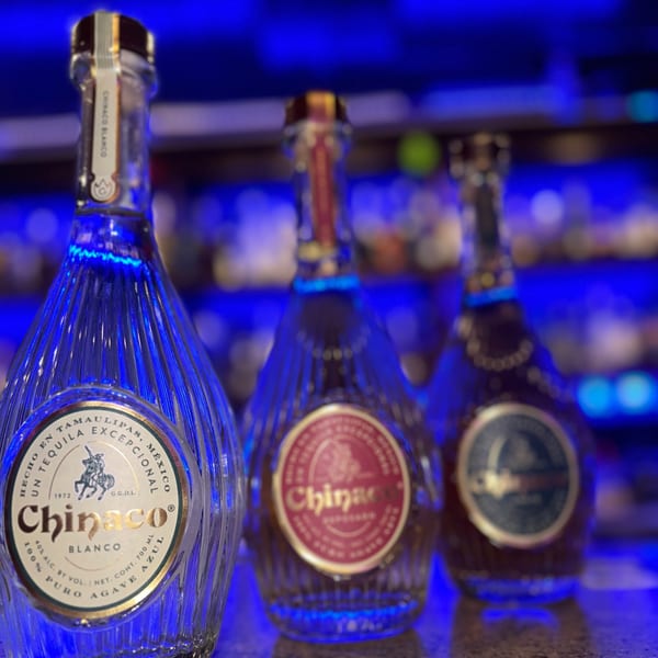Three Chinaco tequila bottles, with the front bottle's label clearly visible, under blue lighting.