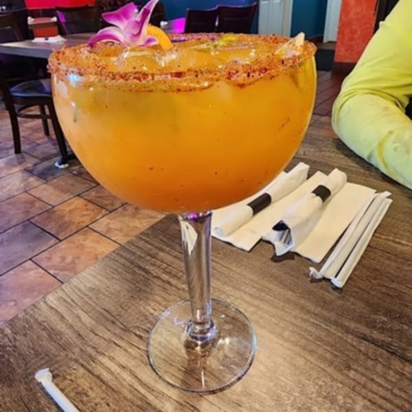 A large orange margarita with a chili-lime rim and orchid garnish sits on a wooden table.