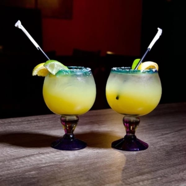 Two vibrant yellow margaritas in goblet glasses with salted rims and lime garnishes on a wooden table.